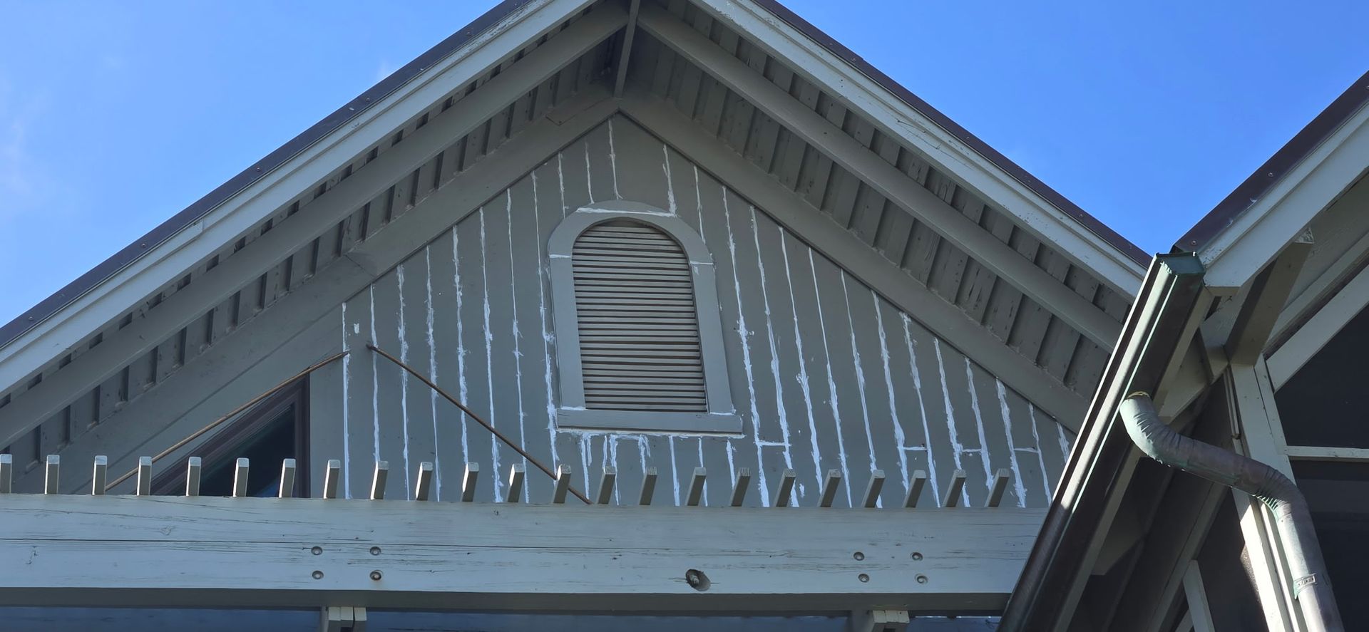 Gray house gable with arched vent and rain gutter against a blue sky.