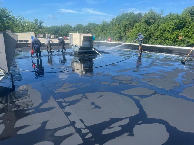 Workers applying sealant to a flat roof, blue sky overhead, trees in the background, sunny day.