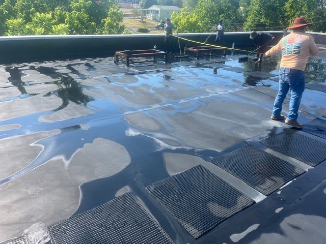 Man on black flat roof, applying sealant. Sunlight reflects on the wet surface.  Yellow caution tape present.