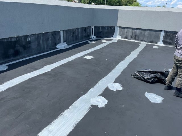 Flat roof with white sealant being applied by a worker, dark surface, gray walls.