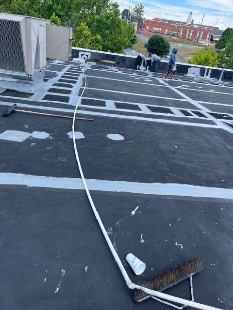 People working on a black flat roof with white paint lines. A long pole and a building are visible.