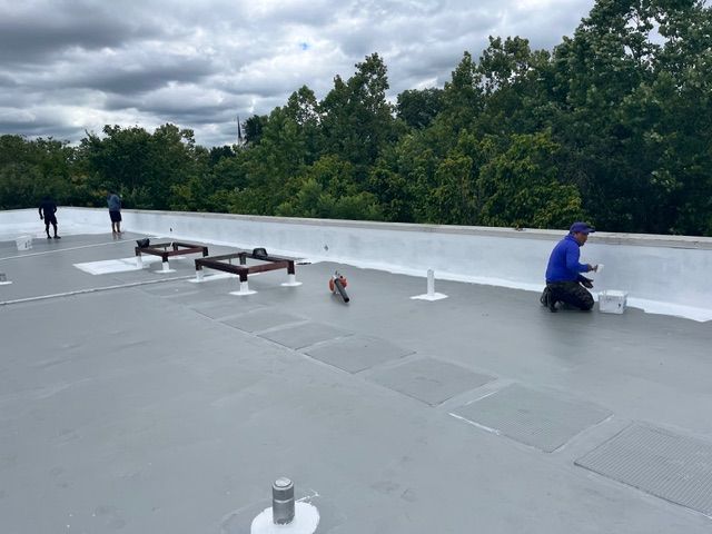 Workers applying white coating to a flat roof, near equipment and trees under a cloudy sky.