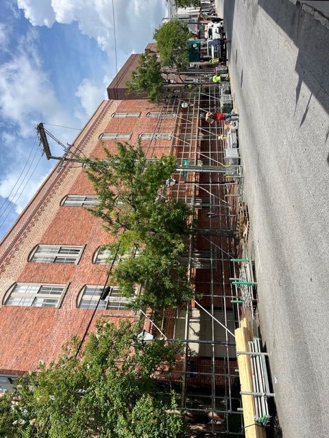 Scaffolding on brick building with trees. Workers visible. Road beside it. Blue sky.
