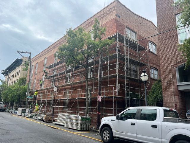 Brick building with scaffolding; workers on the structure. A white truck and supplies are parked nearby.