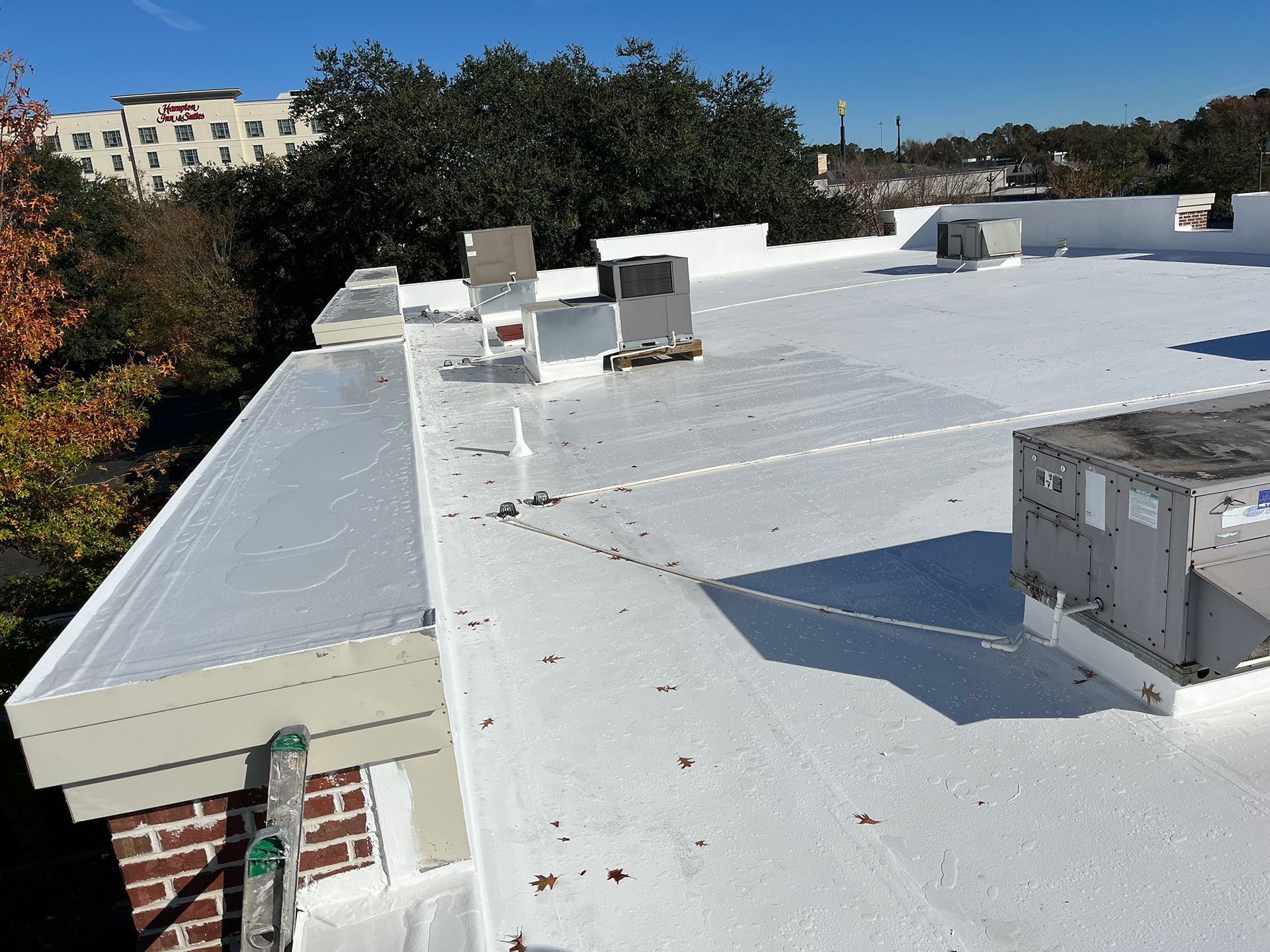 White commercial flat roof with HVAC units and surrounding trees on a sunny day.