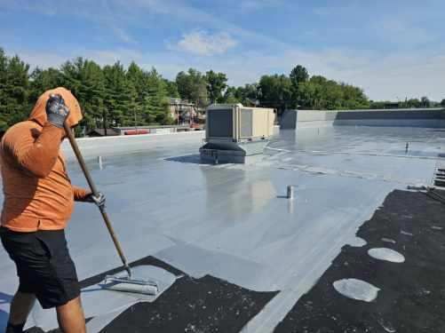 Man applying gray sealant to a flat commercial roof with a roller. Outdoors on a sunny day.