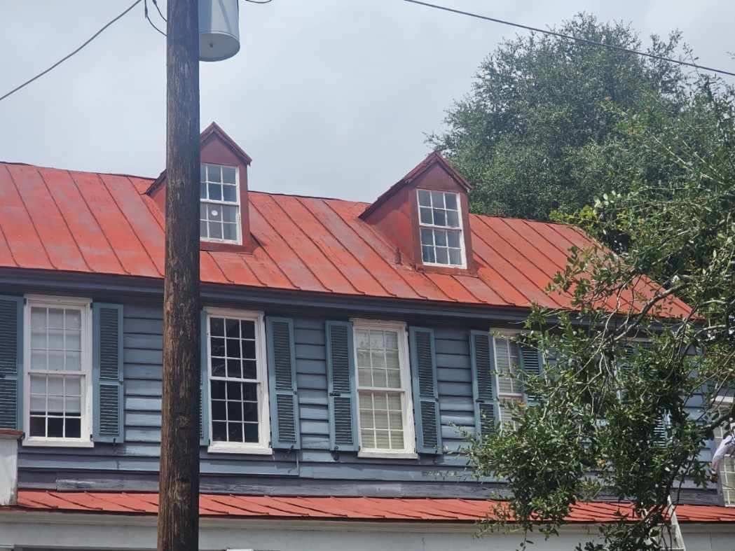 Blue house with red metal roof, two dormers, white trim, shutters, and pole in front.