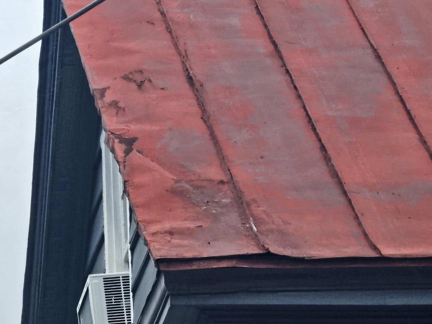 Red metal roof with damage near the edge. Dark building siding and an air conditioning unit are visible.