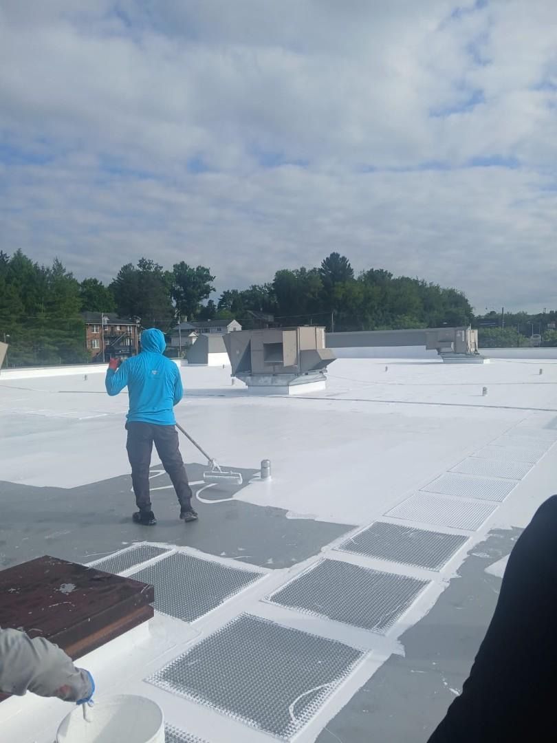 Person in blue jacket applying white coating to a flat roof, sunny day.