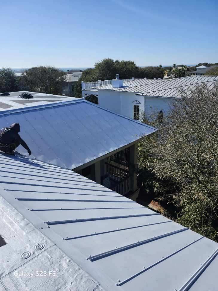 Person on a gray metal roof, possibly working. Several white buildings and trees in the background under a blue sky.