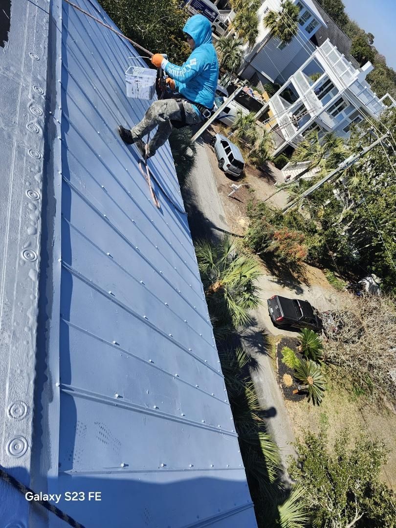 Person on a roof, secured by ropes, painting metal sheets. Green foliage and houses in background.