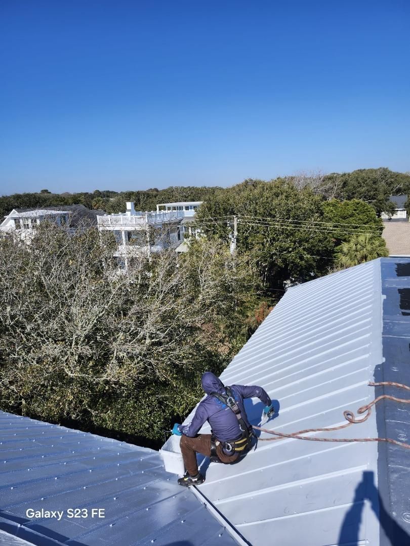 Roofer on a metal roof, secured by a rope. Blue sky, houses, and trees in the background.