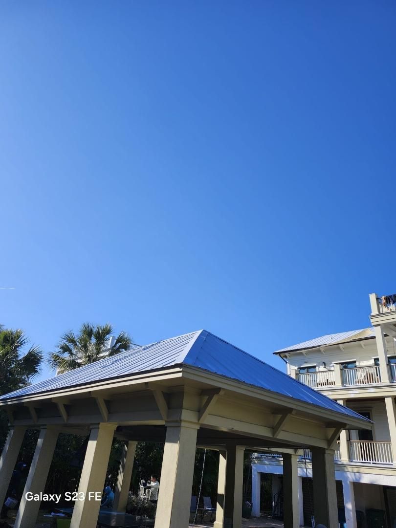 Gazebo with silver roof and beige supports, against a clear blue sky, next to a white building.