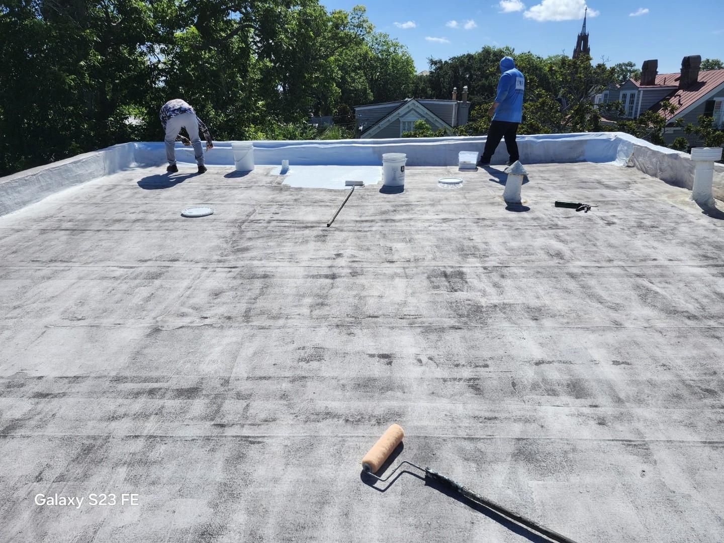 Two workers coating a flat roof with white sealant using rollers, on a sunny day.