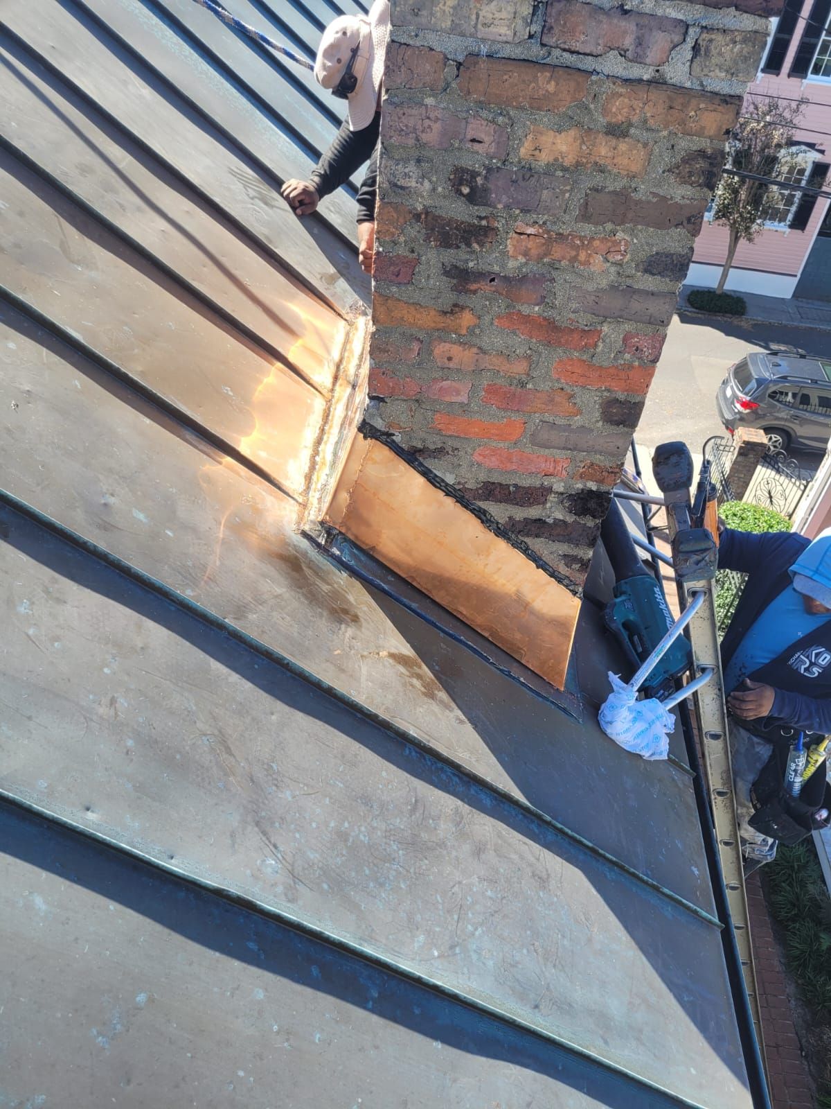 Workers on a roof near a brick chimney; copper flashing around the base; view from above.