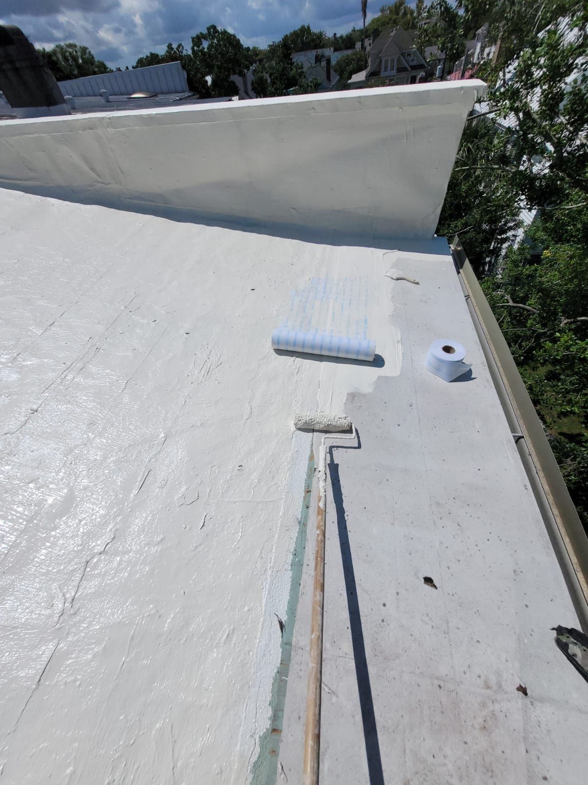 White-coated roof with roller, brush, and sealant; a person appears to be applying a coating on a sunny day.