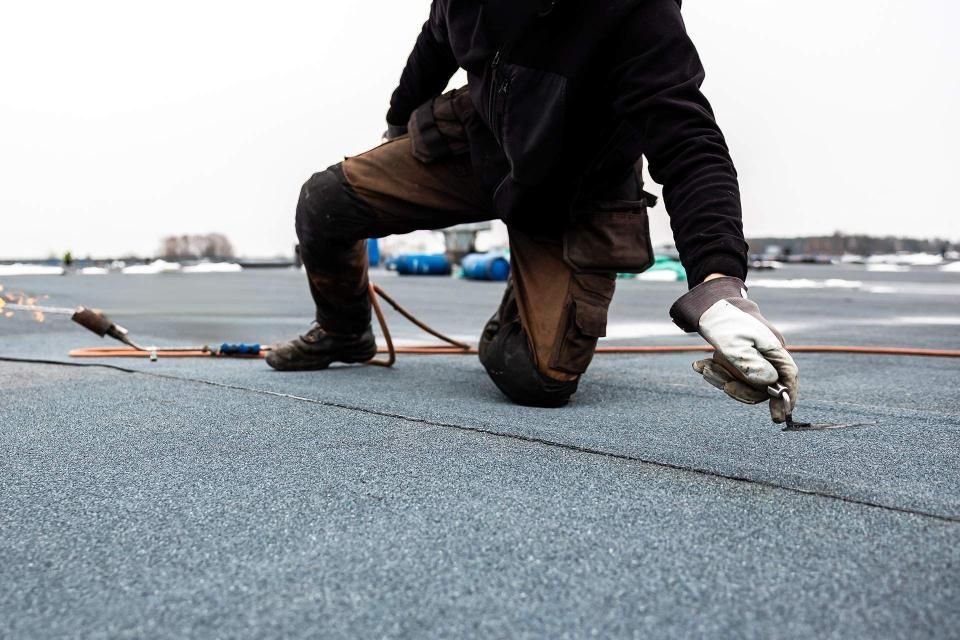 Roofer kneeling on a flat roof, using a torch and tool to seal a seam.