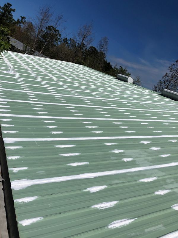 Green metal roof with patches of white substance, trees, and blue sky.