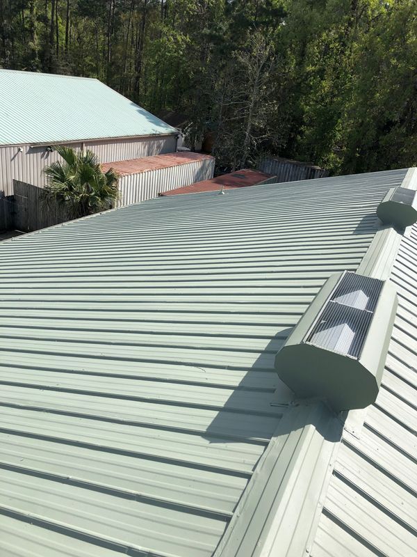 Green metal roof with vents, trees, and buildings in the background on a sunny day.