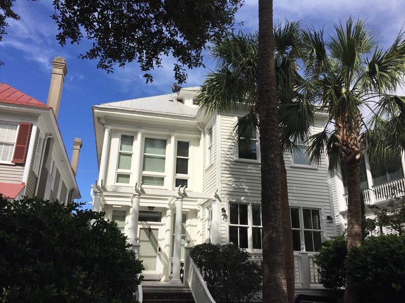 White house with stairs, windows, and a chimney, framed by palm trees and blue sky.