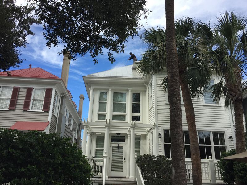 White house with large windows, palm trees, and a red-roofed building on the left under a blue sky.