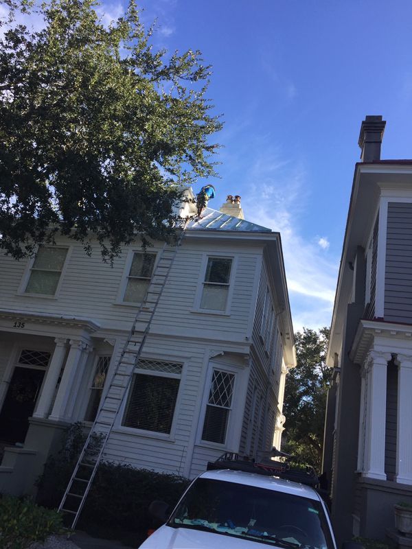 White house with ladder on the side, next to a gray building, tree in the corner, blue sky.