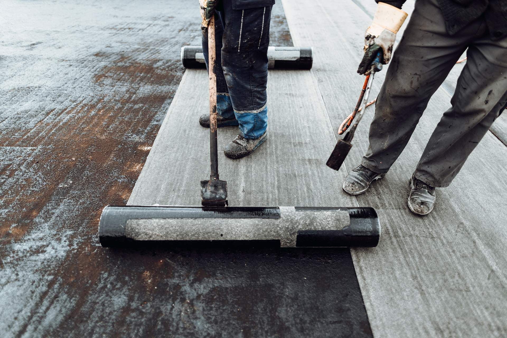 Two workers installing roofing material, one using a roller, the other with a torch.