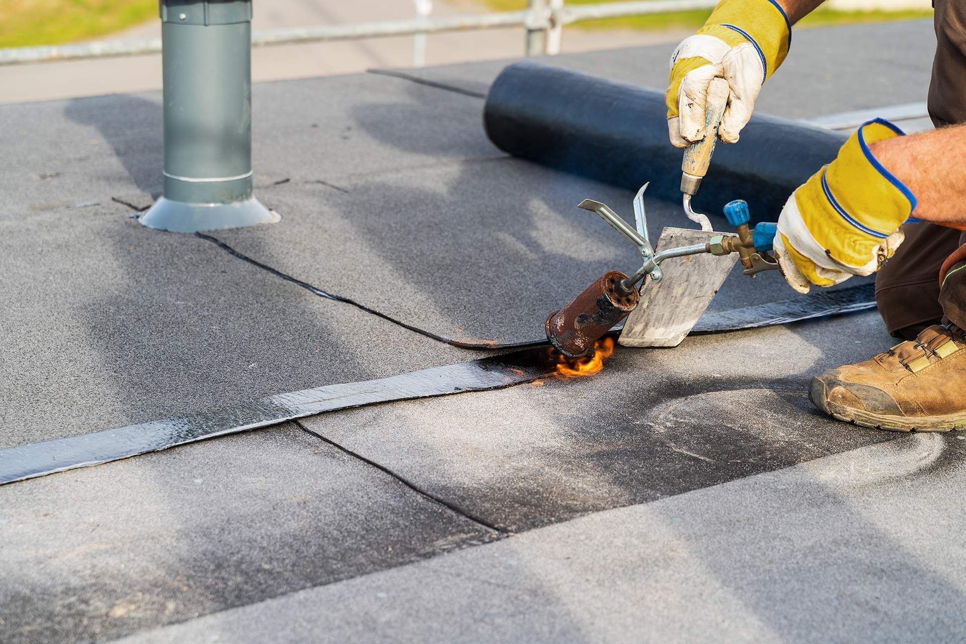 A worker in yellow gloves welds a seam on a flat roof with a torch.