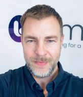Man with short, graying hair and beard smiling, wearing a dark shirt in front of a logo.