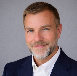 Man with short, graying hair and beard smiling, wearing a dark shirt in front of a logo.
