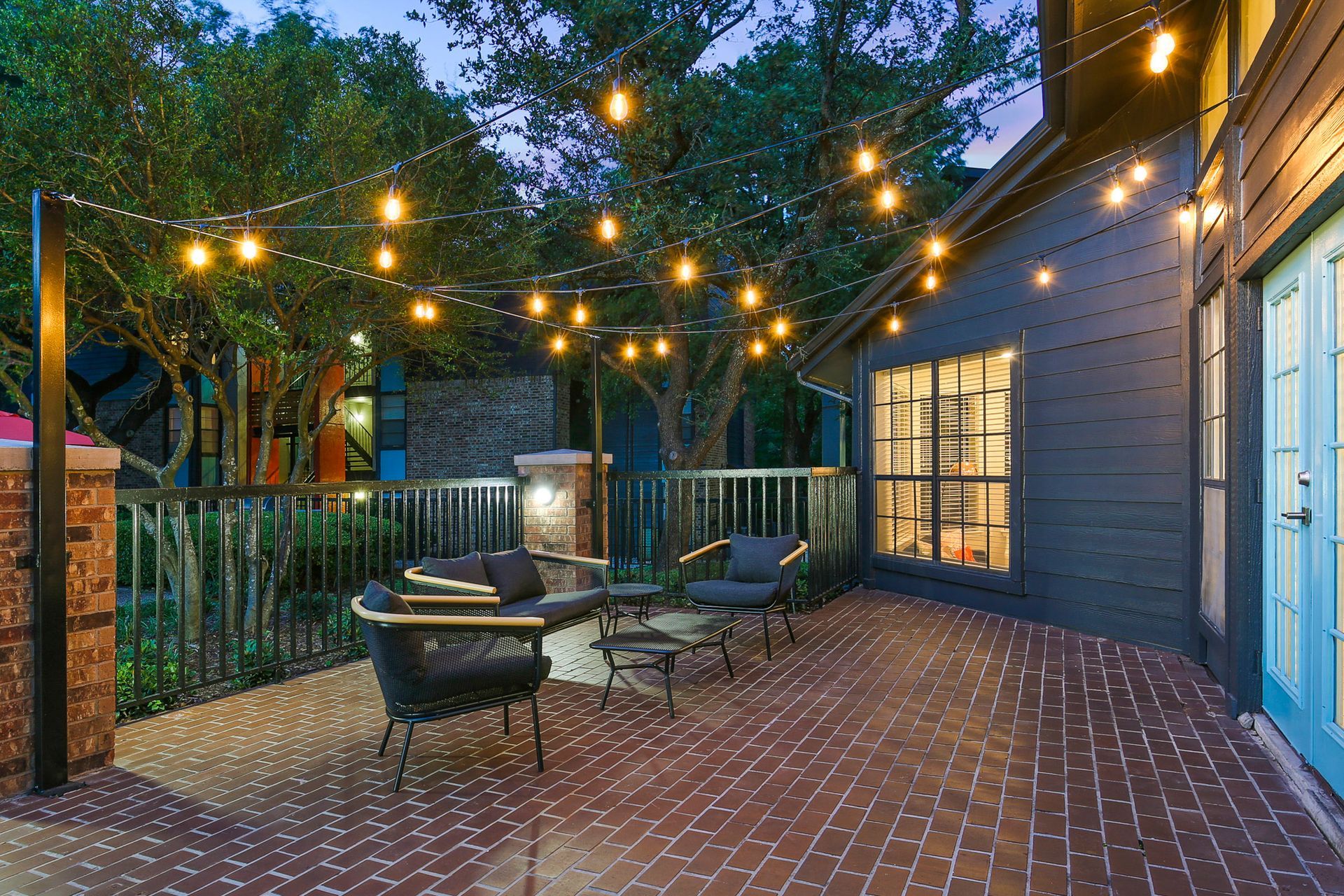 Patio with string lights, brick floor, black furniture, and a blue house at dusk.