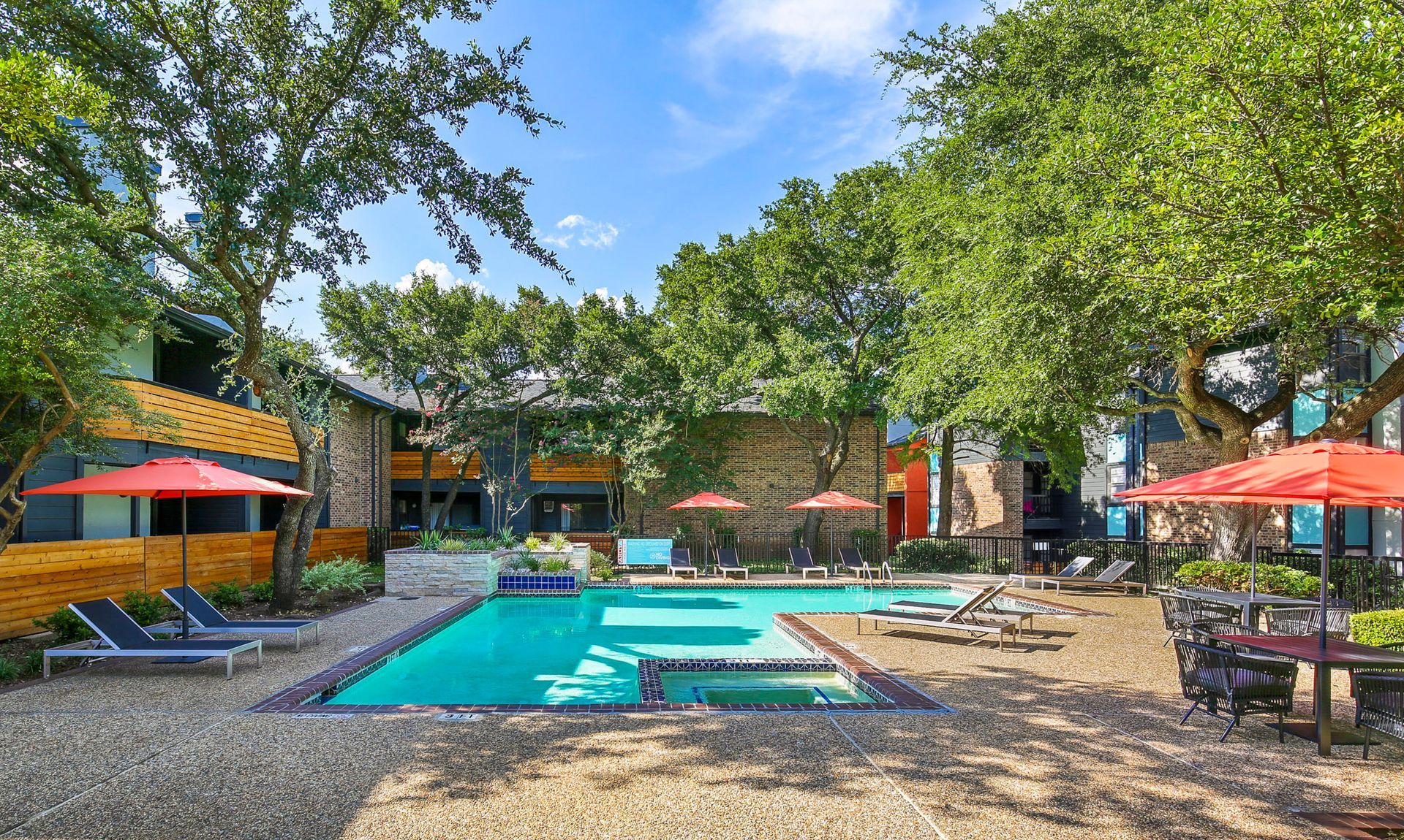 Swimming pool surrounded by lounge chairs, trees, and buildings, under a blue sky.