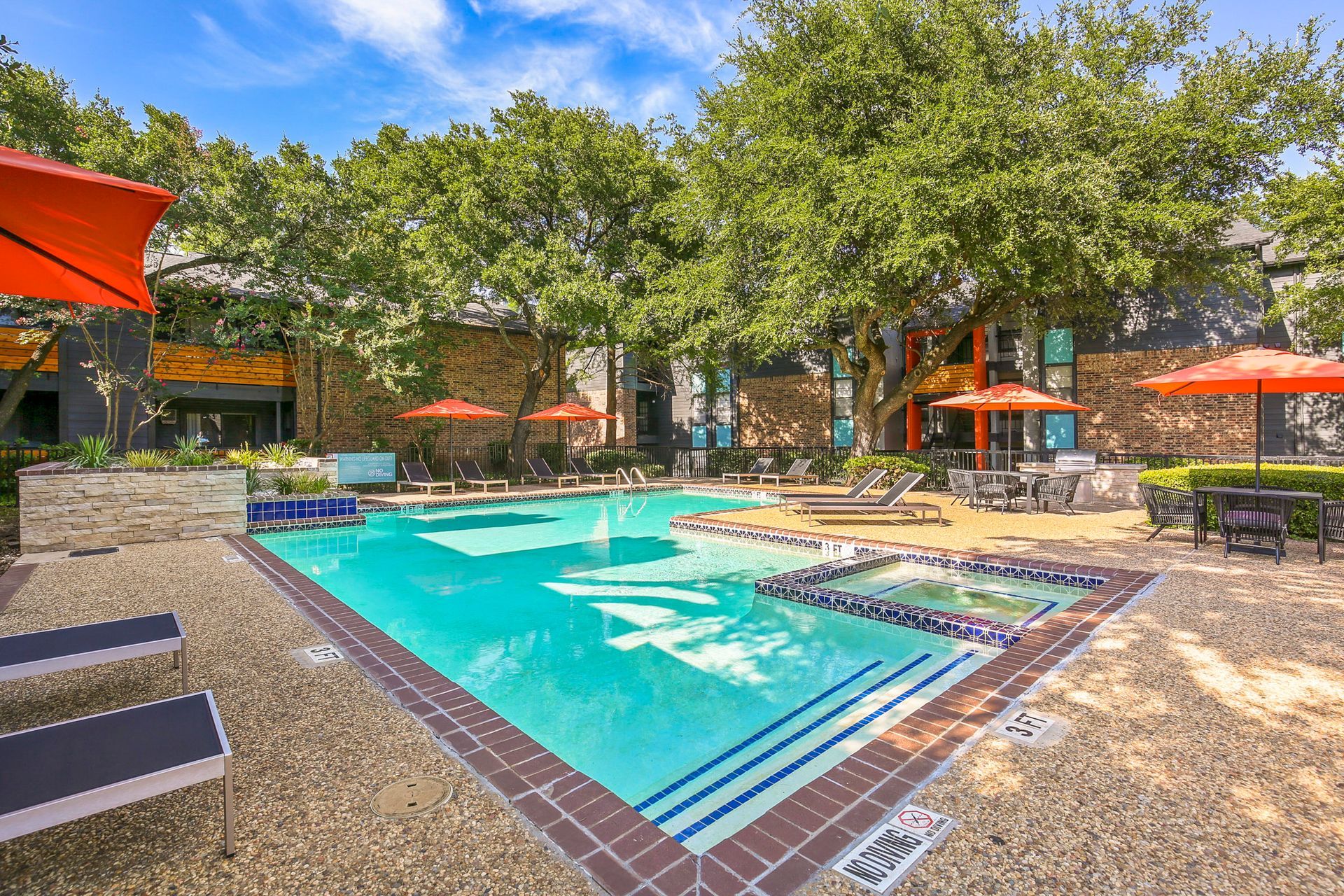 Pool with lounge chairs and orange umbrellas, surrounded by trees and buildings.