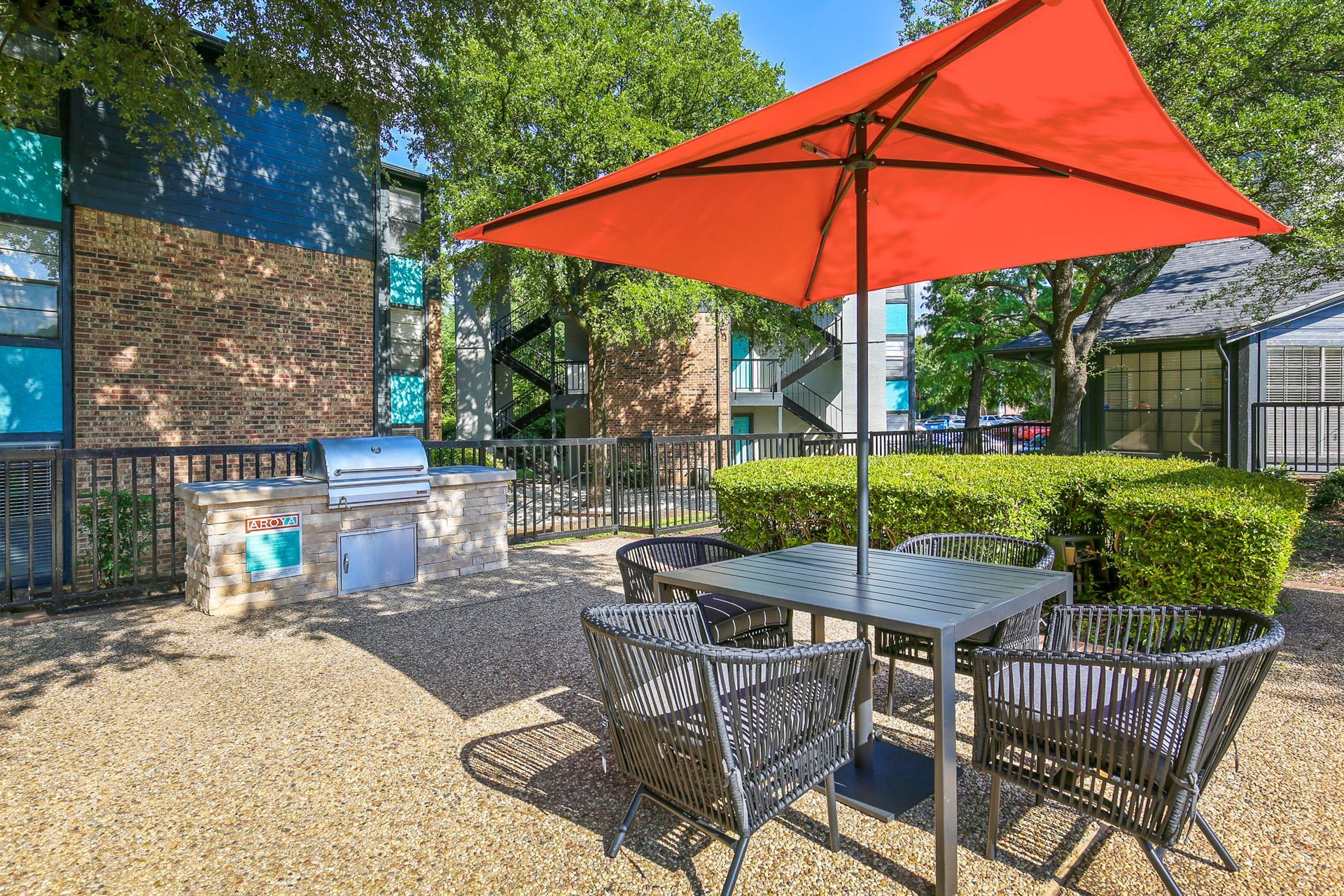 Outdoor seating area with grill, orange umbrella, and turquoise and brick building background.