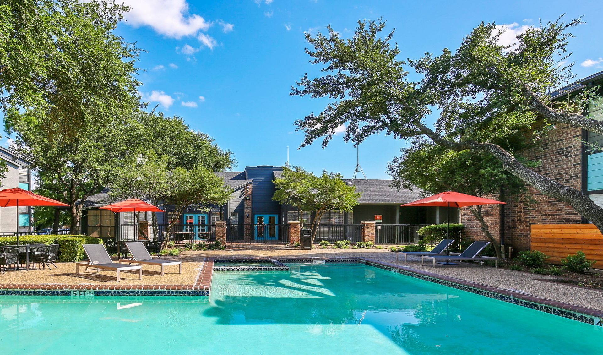 Pool with lounge chairs, orange umbrellas, and building with blue doors under a sunny sky.