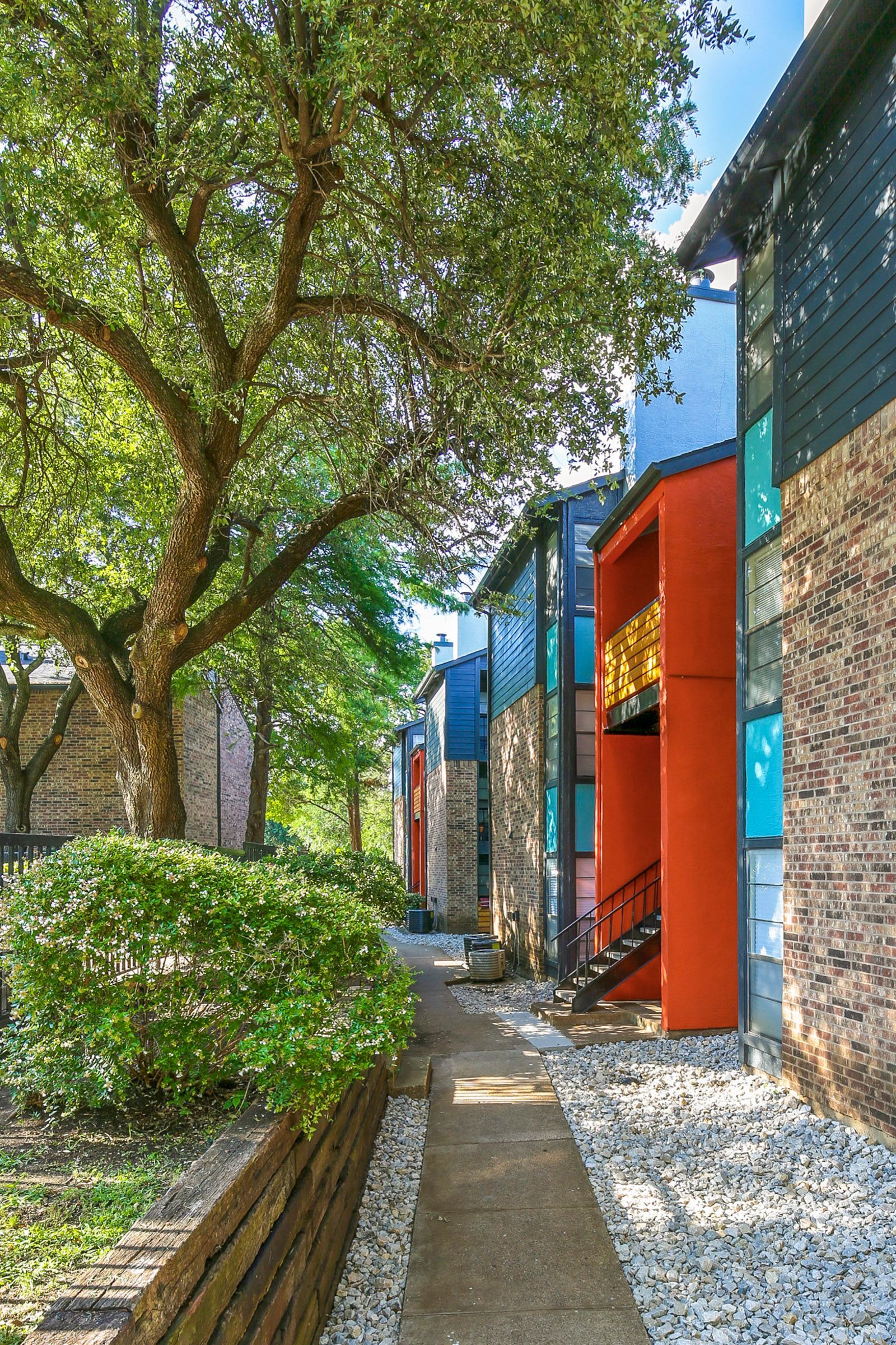 Apartment buildings with colorful accents and a walkway lined with white stones, under a large tree.