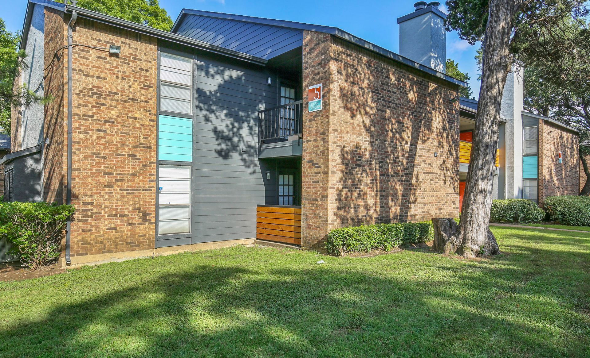 Brick apartment building with black and gray accents, lush lawn, and large tree.