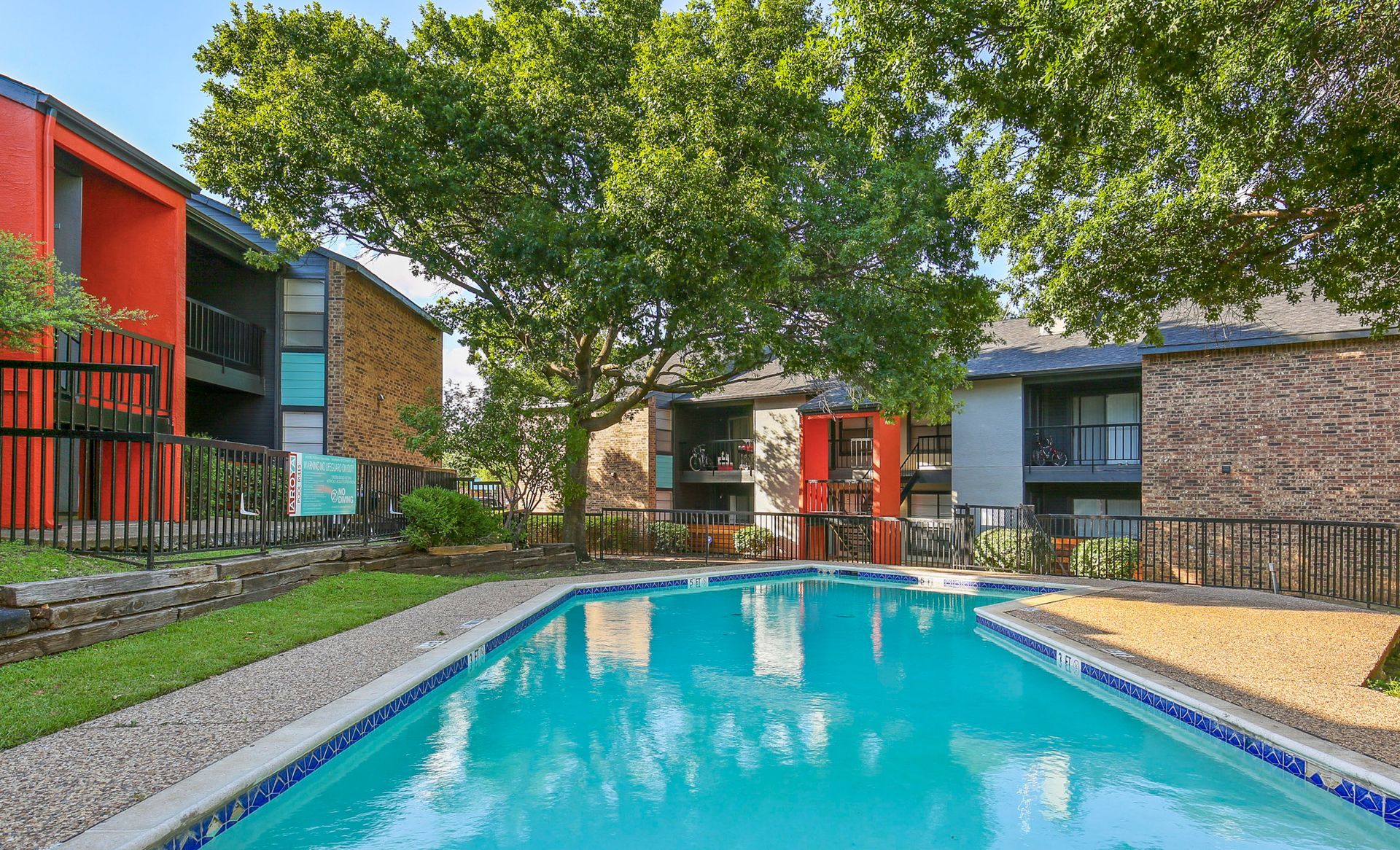 Apartment complex with a pool; turquoise water, colorful buildings, and a large tree.