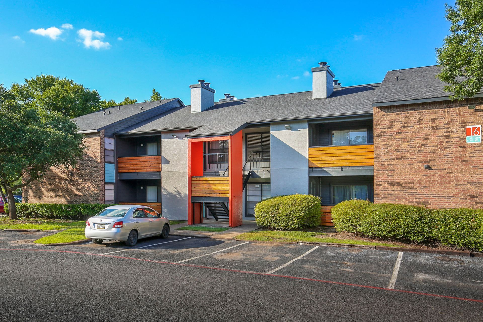 Apartment building exterior, tan car in front. Blue sky, orange and wood accents, parking area.