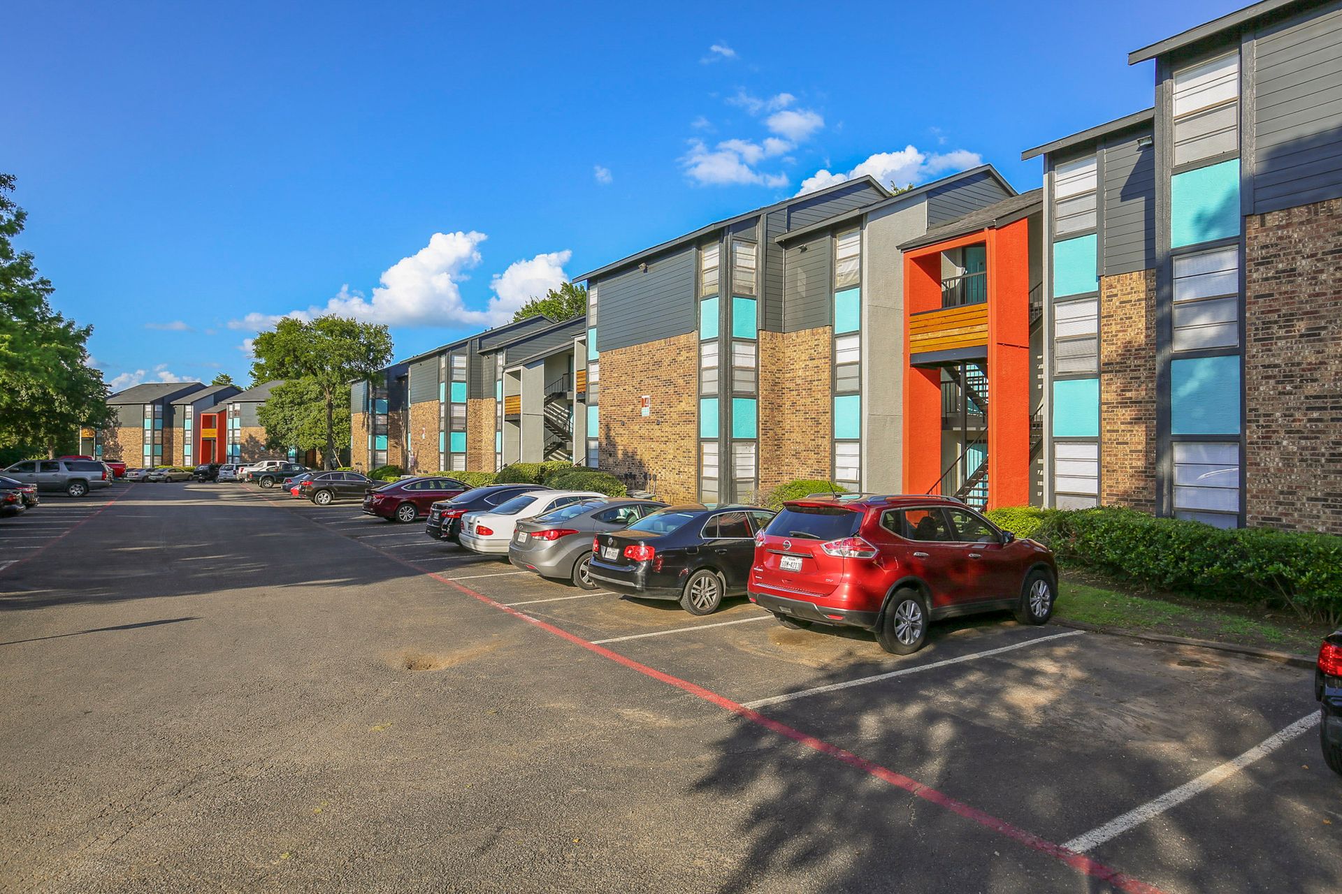 Apartment complex with parked cars under a blue sky. Buildings feature brick, teal, gray, and orange accents.