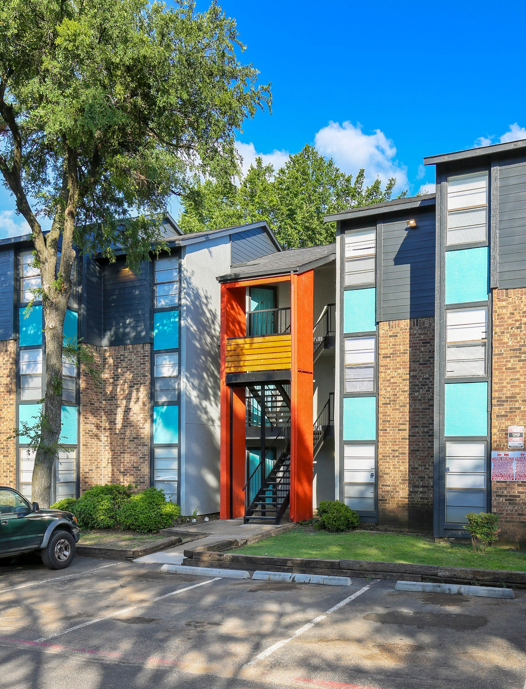 Apartment building exterior, brick and teal accents, orange entrance, parked car, green tree.