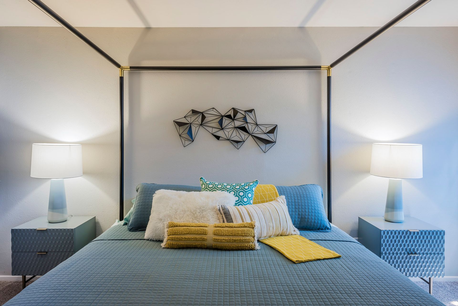 Bedroom with canopy bed, nightstands with lamps, pillows, blue and yellow bedding, and wall art.