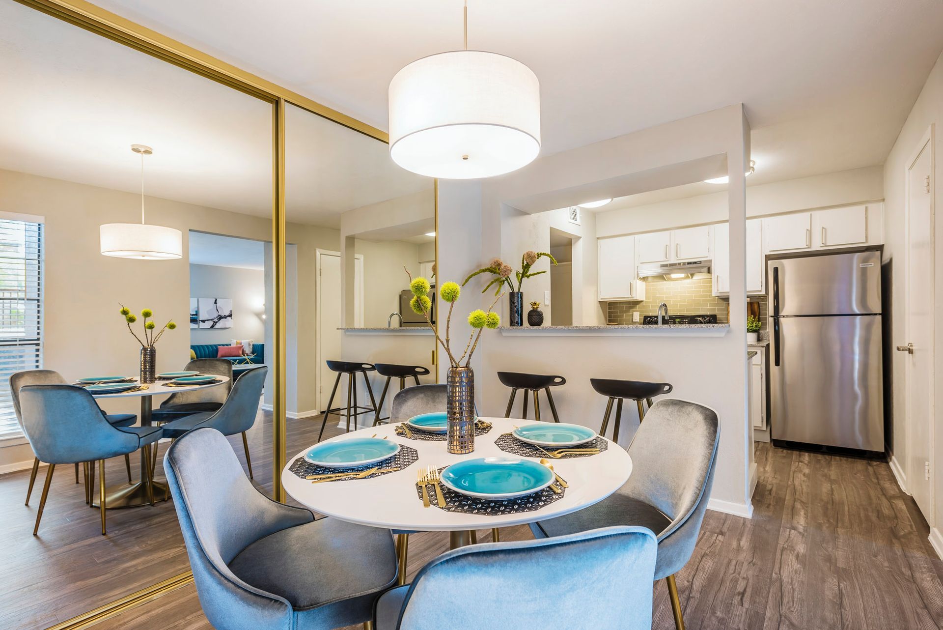 Dining area with round table, blue chairs, mirrored wall, and open kitchen with stainless steel appliances.