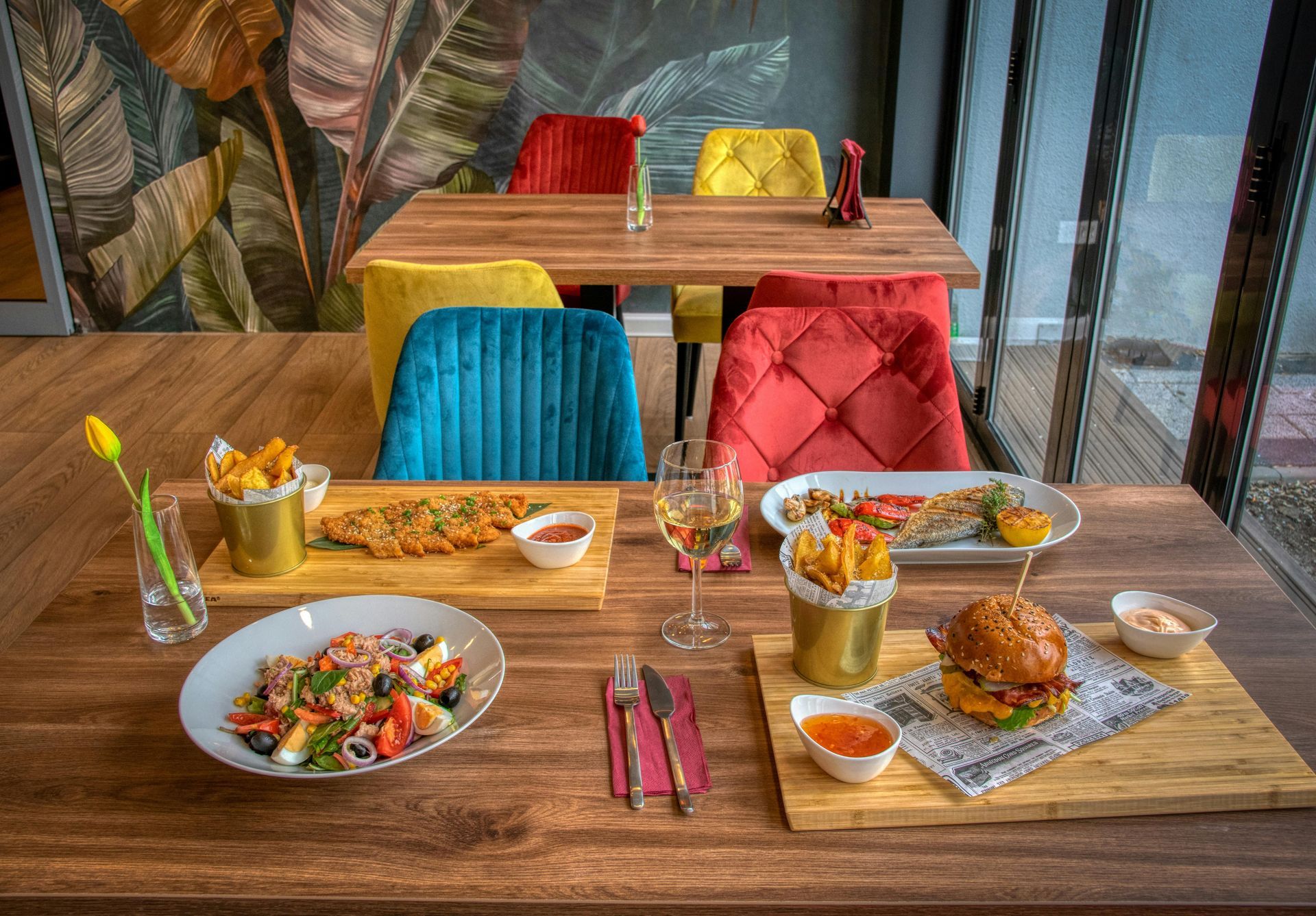 A table set for a meal with colorful chairs. Dishes include a salad, burger, fries, and drinks.