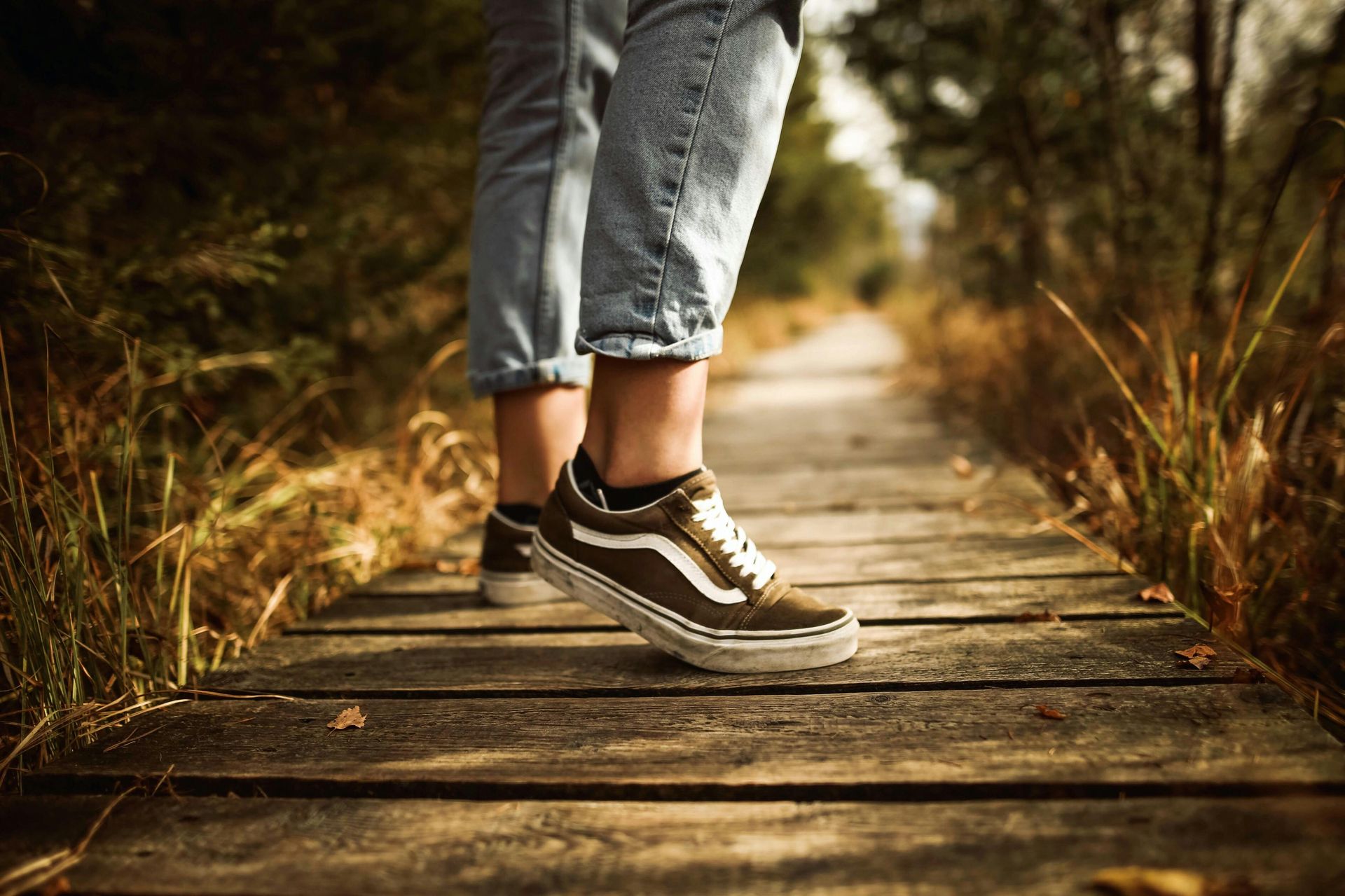Person's legs and feet wearing sneakers on a wooden walkway in a natural setting, with tall grass.