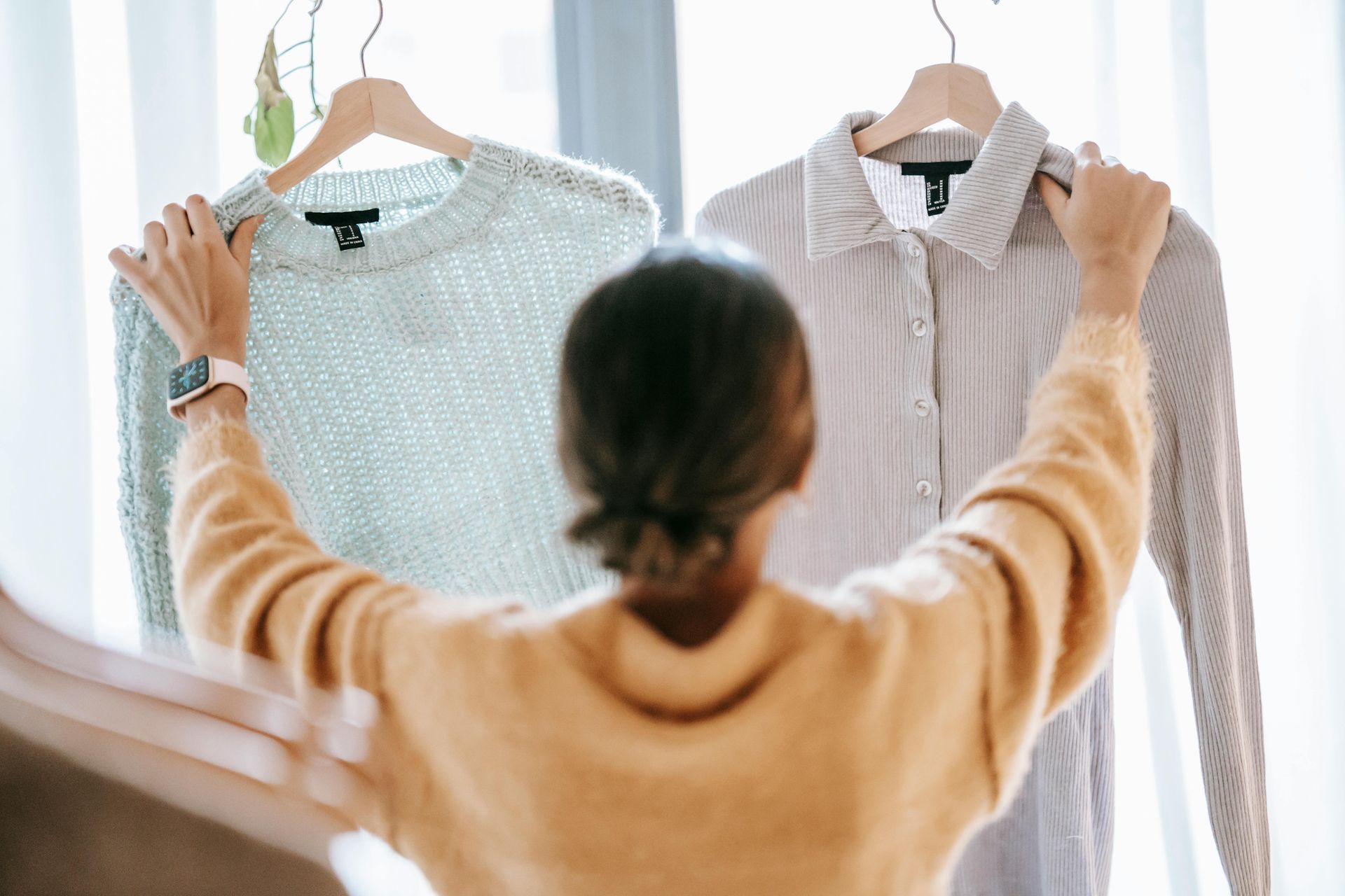 Person holding up a light blue sweater and a button-down shirt on hangers.