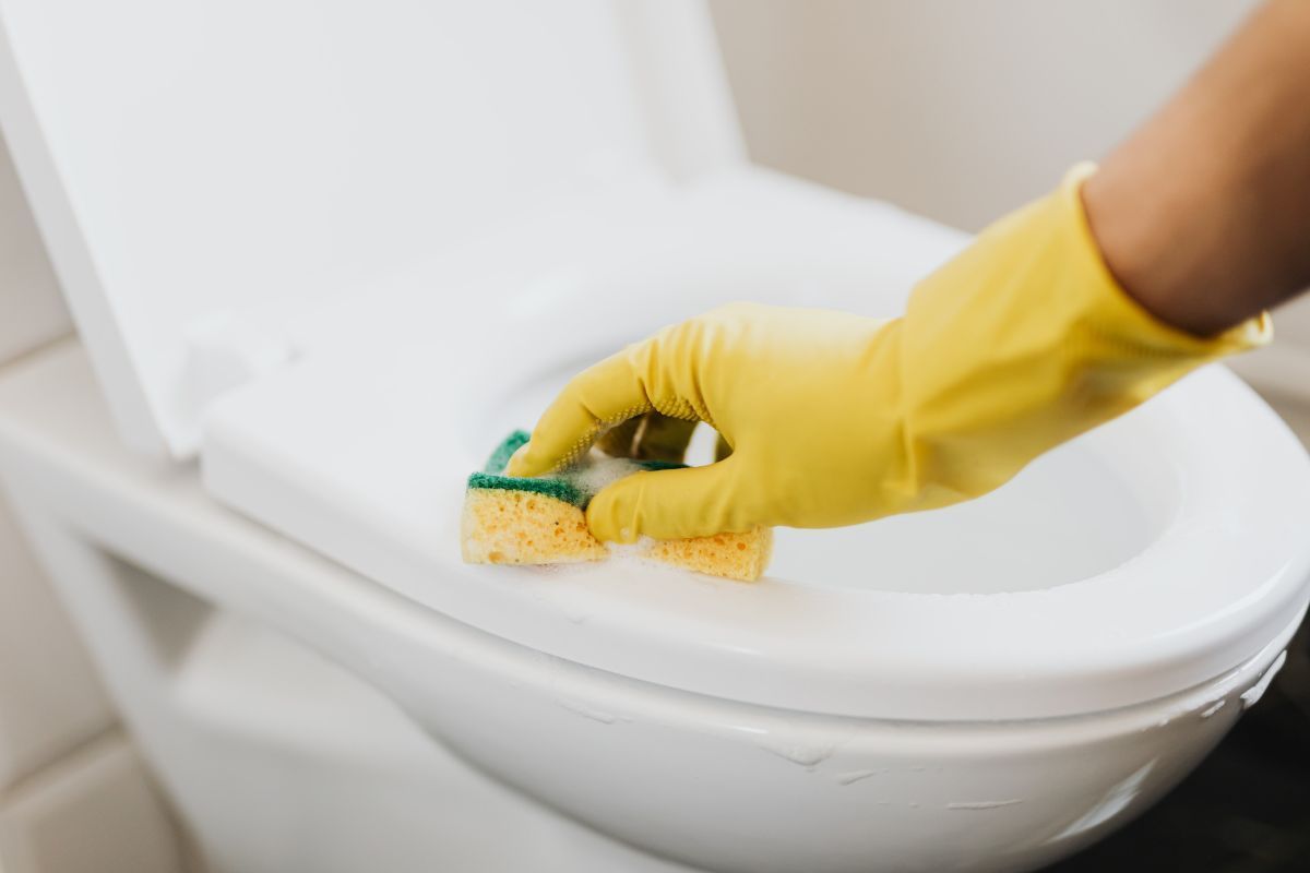 A person wearing yellow gloves is cleaning a toilet with a sponge.