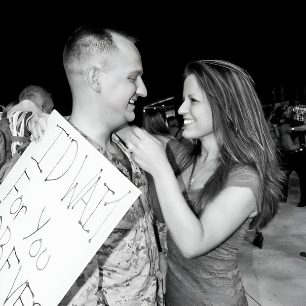 Smiling couple outdoors at night, woman adjusting the man's shirt while he holds a sign.