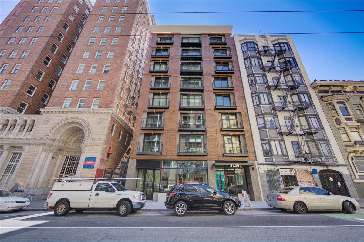 Buildings on a city street; brick building in the center, flanked by older structures and parked cars.