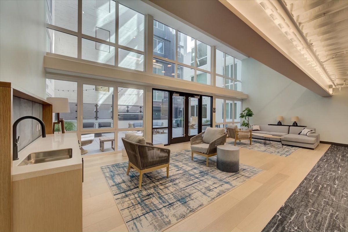 Lobby with large windows, seating area with rug, light wood floors, and a kitchen area.
